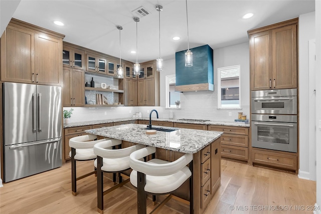 kitchen featuring appliances with stainless steel finishes, a kitchen island with sink, wall chimney range hood, sink, and light hardwood / wood-style flooring