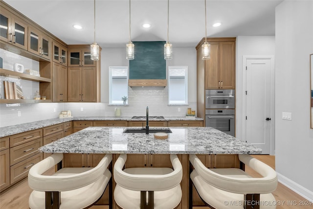 kitchen with hanging light fixtures, tasteful backsplash, light stone counters, double oven, and a center island with sink