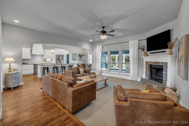 living room with ceiling fan with notable chandelier, light wood-type flooring, and a stone fireplace