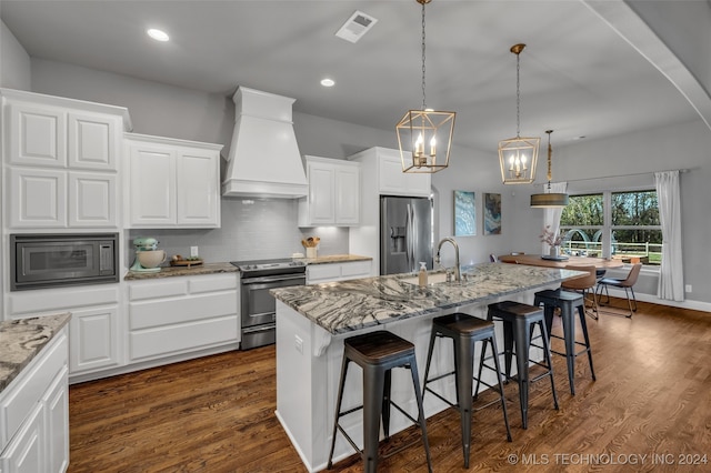 kitchen with custom exhaust hood, a center island with sink, appliances with stainless steel finishes, hanging light fixtures, and white cabinetry