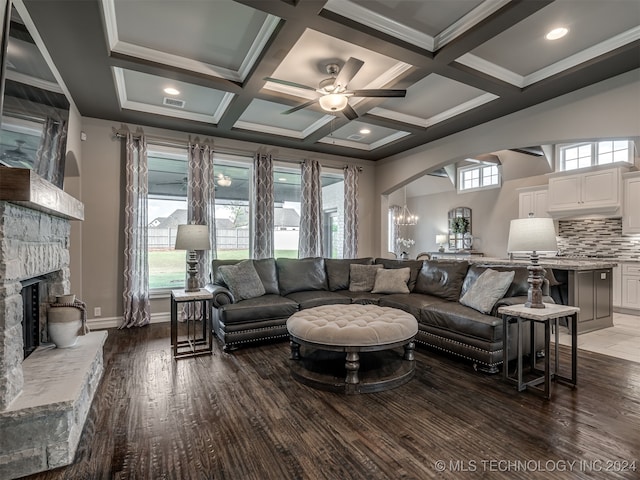 living room with a stone fireplace, ceiling fan with notable chandelier, coffered ceiling, dark hardwood / wood-style floors, and beam ceiling