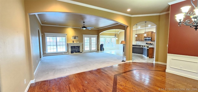 unfurnished living room featuring light wood finished floors, arched walkways, a glass covered fireplace, crown molding, and ceiling fan with notable chandelier