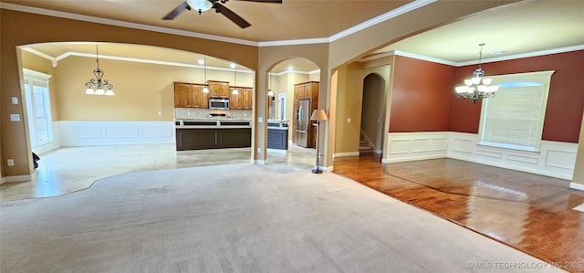 unfurnished living room with arched walkways, wainscoting, light carpet, and ceiling fan with notable chandelier