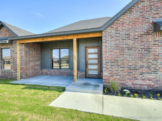 doorway to property with a shingled roof, a yard, brick siding, and a patio