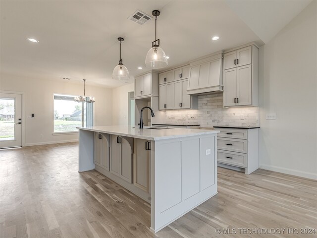 kitchen featuring hanging light fixtures, sink, premium range hood, light wood-type flooring, and a kitchen island with sink