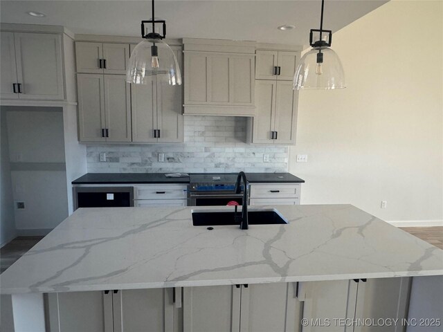 kitchen with decorative light fixtures, sink, stainless steel stove, light stone counters, and a kitchen island with sink