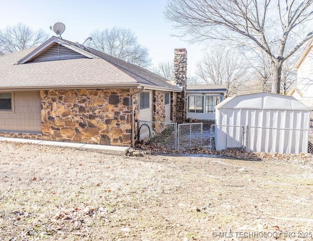back of house featuring a shingled roof, stone siding, fence, a chimney, and a storage shed