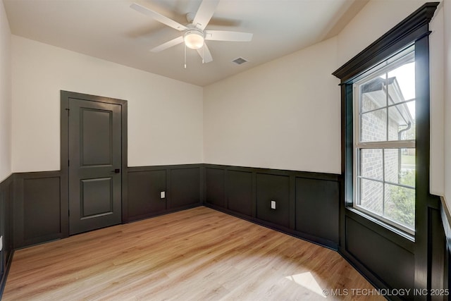 empty room with wainscoting, light wood-type flooring, visible vents, ceiling fan, and a decorative wall