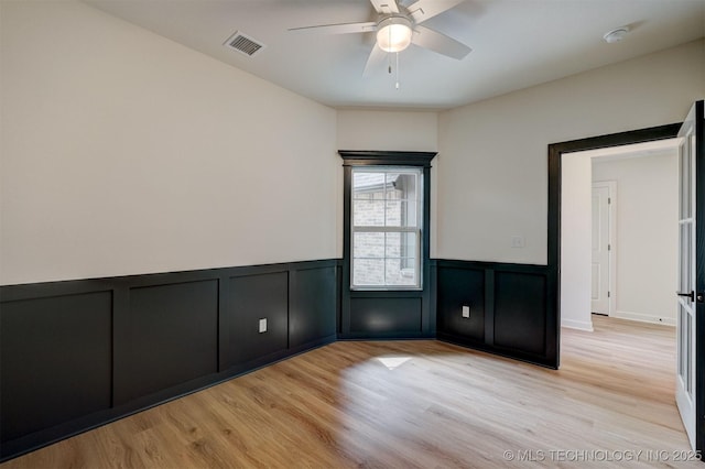 empty room featuring ceiling fan, light wood-style flooring, visible vents, and a wainscoted wall