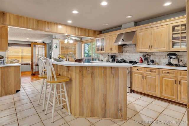 kitchen with an island with sink, stainless steel gas range oven, wall chimney exhaust hood, and light tile patterned flooring