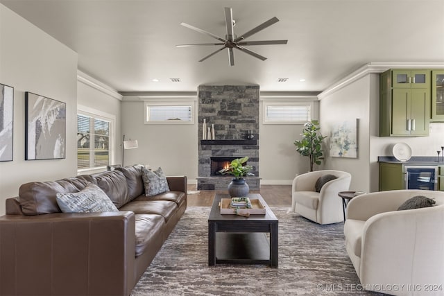 living room with beverage cooler, dark wood-type flooring, ceiling fan, crown molding, and a stone fireplace