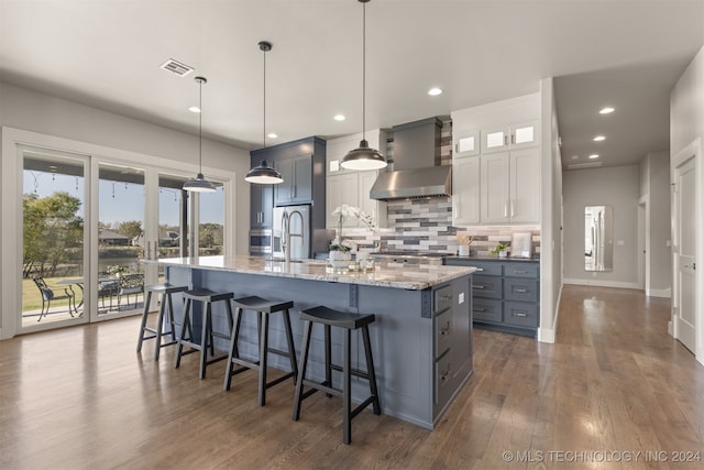 kitchen featuring white cabinets, pendant lighting, wall chimney range hood, and an island with sink