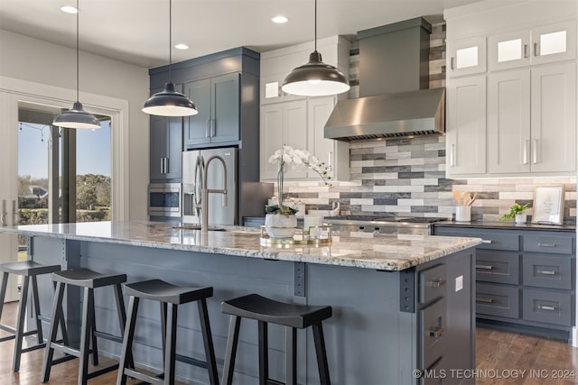 kitchen with appliances with stainless steel finishes, pendant lighting, a kitchen island with sink, and wall chimney exhaust hood