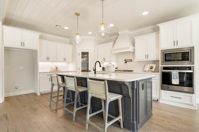 kitchen featuring stainless steel appliances, a center island with sink, custom range hood, white cabinetry, and light hardwood / wood-style flooring