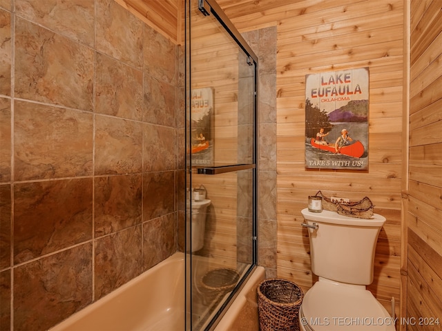 bathroom featuring bath / shower combo with glass door, wooden walls, and toilet