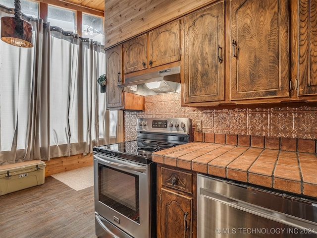 kitchen with tile counters, wood finished floors, stainless steel appliances, under cabinet range hood, and backsplash