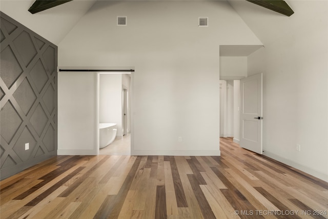 unfurnished bedroom with ensuite bathroom, a barn door, high vaulted ceiling, and light hardwood / wood-style flooring