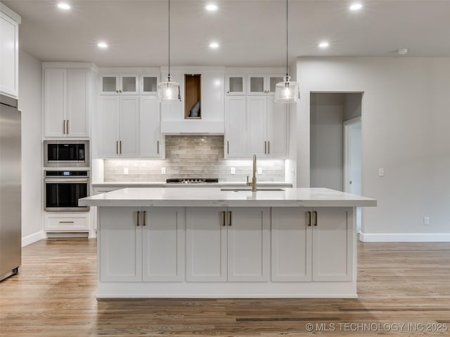 kitchen with a sink, an island with sink, built in appliances, premium range hood, and white cabinetry