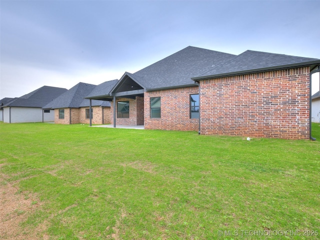back of property with a lawn, a shingled roof, brick siding, and a patio
