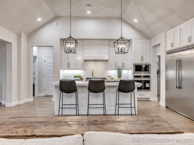 kitchen with an island with sink, built in appliances, white cabinets, and decorative light fixtures