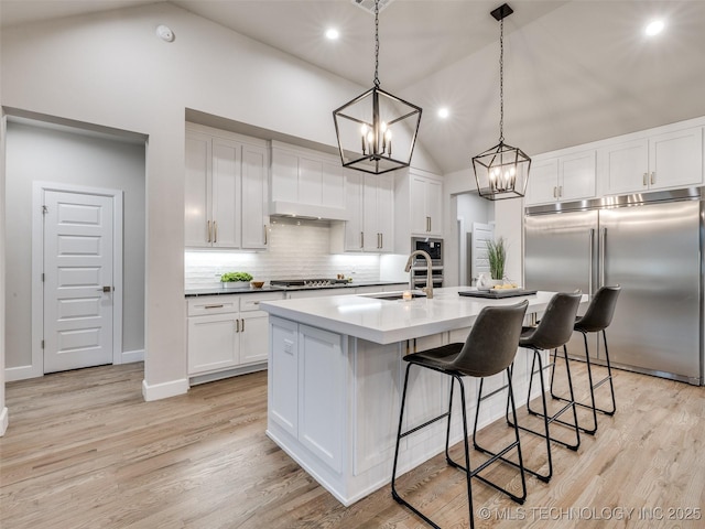 kitchen with white cabinetry, built in appliances, hanging light fixtures, and a center island with sink