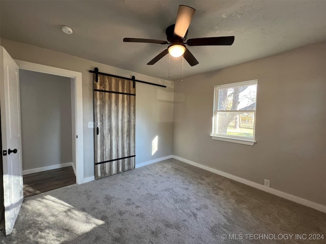 unfurnished bedroom with a barn door, dark carpet, a ceiling fan, and baseboards