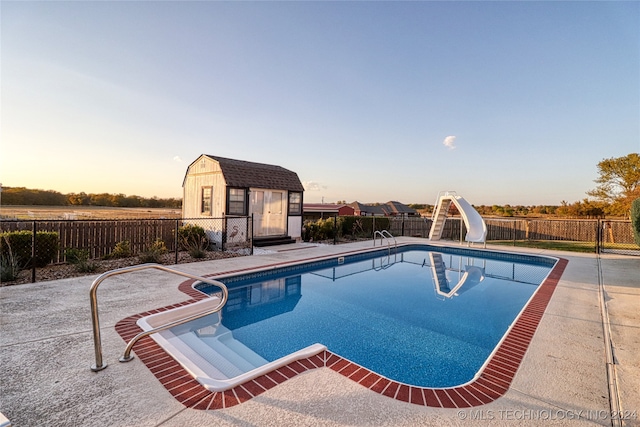 pool at dusk with a patio area, a water slide, and a storage shed