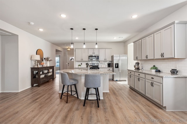 kitchen featuring light stone countertops, pendant lighting, a center island with sink, appliances with stainless steel finishes, and light wood-type flooring