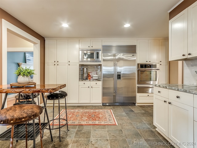 kitchen with white cabinets, built in appliances, dark stone counters, and backsplash