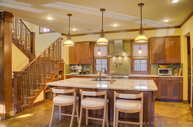 kitchen featuring a kitchen island with sink, backsplash, sink, wall chimney exhaust hood, and decorative light fixtures