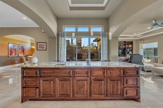 kitchen with light stone counters, ornamental molding, a tray ceiling, a kitchen island with sink, and ceiling fan