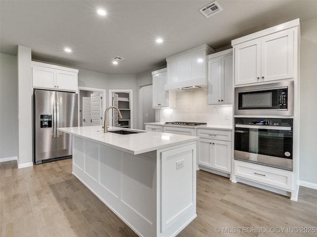 kitchen with a sink, a kitchen island with sink, custom range hood, appliances with stainless steel finishes, and visible vents