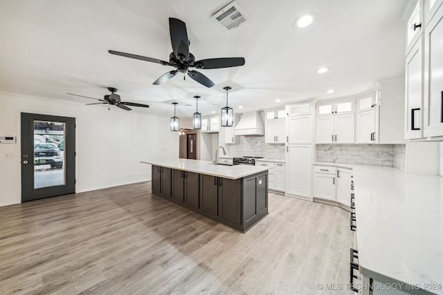 kitchen with custom range hood, white cabinets, light hardwood / wood-style flooring, pendant lighting, and a center island with sink