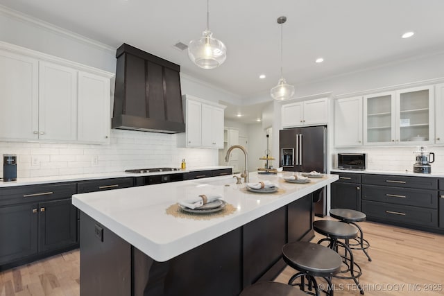 kitchen featuring high end fridge, white cabinetry, an island with sink, and wall chimney exhaust hood