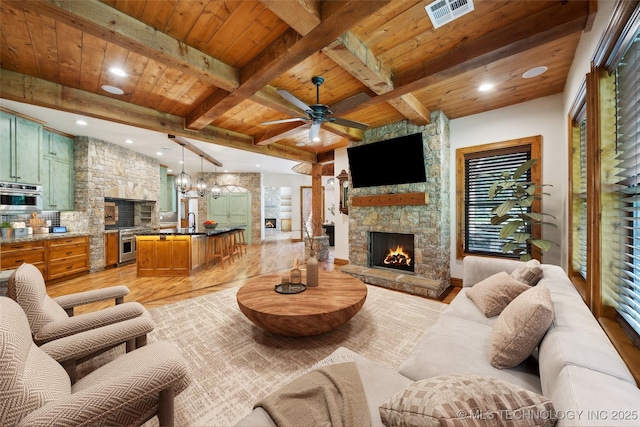 living room with ceiling fan with notable chandelier, a stone fireplace, light hardwood / wood-style floors, wooden ceiling, and beam ceiling