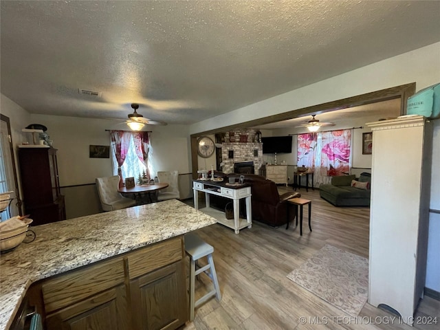 kitchen with a brick fireplace, ceiling fan, light stone countertops, a textured ceiling, and light hardwood / wood-style floors