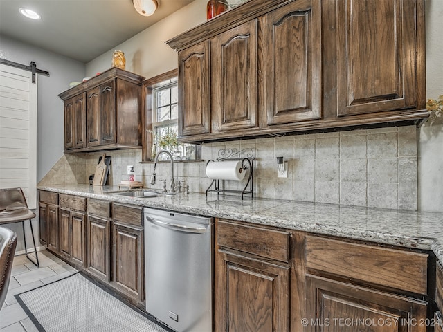 kitchen featuring sink, stainless steel dishwasher, decorative backsplash, a barn door, and light stone counters