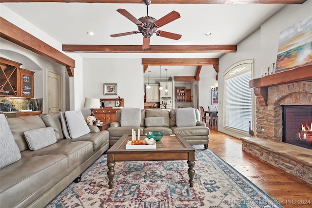 living room with beamed ceiling, ceiling fan, a stone fireplace, and light wood-type flooring