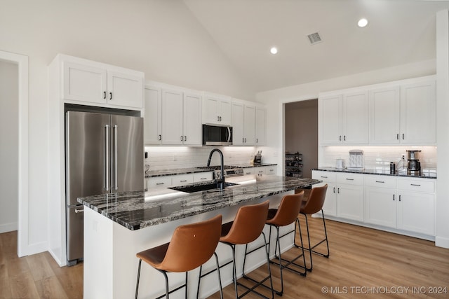 kitchen with decorative backsplash, appliances with stainless steel finishes, a kitchen island with sink, dark stone countertops, and white cabinets