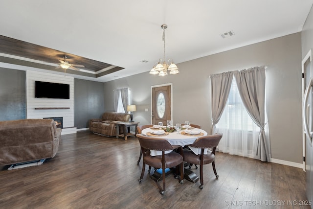 dining space featuring a large fireplace, a tray ceiling, dark hardwood / wood-style flooring, and ceiling fan with notable chandelier