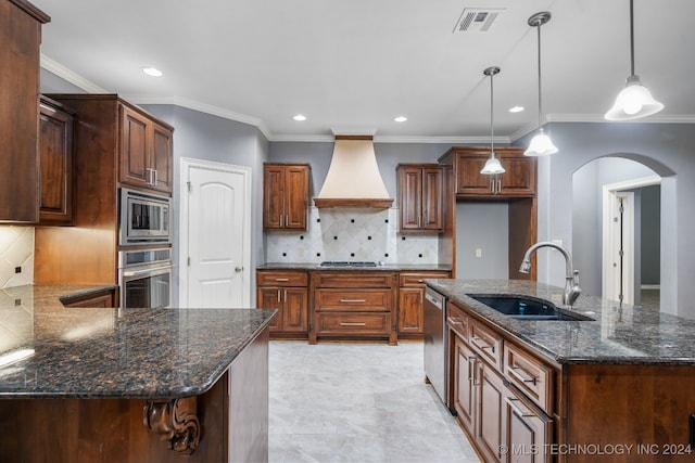 kitchen with custom range hood, stainless steel appliances, sink, decorative light fixtures, and a center island with sink