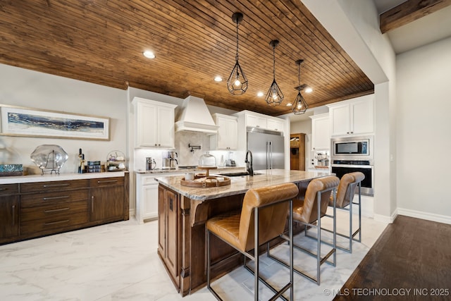 kitchen with built in appliances, premium range hood, a kitchen island with sink, backsplash, and white cabinetry