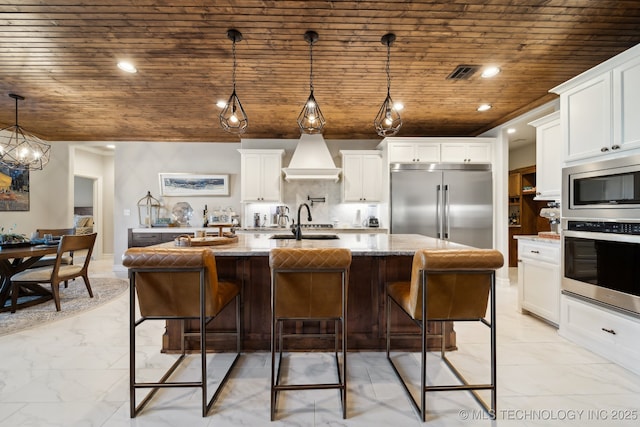 kitchen with built in appliances, pendant lighting, a center island with sink, stone countertops, and white cabinetry