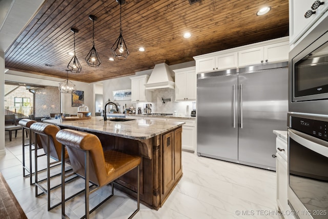 kitchen with built in appliances, decorative light fixtures, white cabinets, premium range hood, and a kitchen island with sink