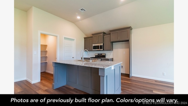 kitchen with lofted ceiling, backsplash, a center island with sink, light stone countertops, and appliances with stainless steel finishes