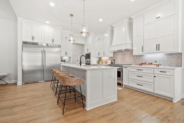 kitchen featuring hanging light fixtures, built in appliances, custom range hood, white cabinets, and a center island with sink