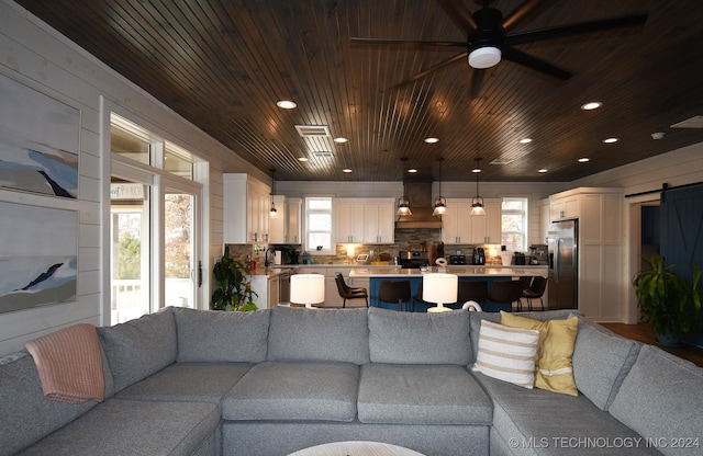 living room with a barn door, wooden ceiling, ceiling fan, and wooden walls