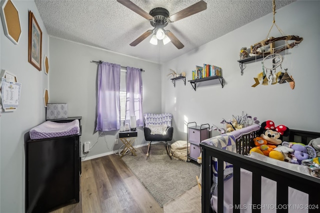 bedroom with wood-type flooring, a nursery area, a textured ceiling, and ceiling fan