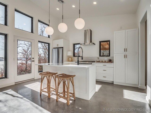 kitchen with wall chimney exhaust hood, stainless steel fridge, decorative light fixtures, a center island with sink, and white cabinets