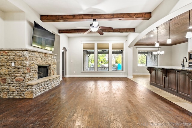 unfurnished living room featuring sink, beamed ceiling, dark hardwood / wood-style floors, a fireplace, and ceiling fan with notable chandelier